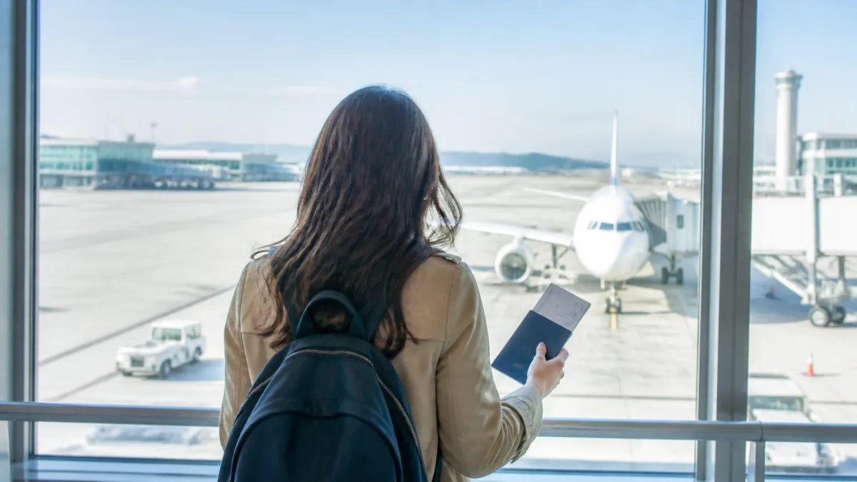 Traveler holding a passport and itinerary while looking at an aircraft through an airport window